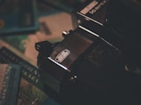 A close-up view of an old-fashioned camera with the branding 'Zeiss Ikon Voigtländer' on top. The camera sits on top of various colorful papers, possibly currency or travel documents, indicating a vintage or travel theme.