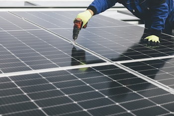 A person wearing gloves and a blue protective suit is using a power drill on a large solar panel array, indicating maintenance or installation work.