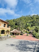 A scenic view of a picturesque estate with a large rustic house on the left, surrounded by lush greenery and trees. The bright blue sky with white clouds adds a vibrant backdrop. In the middle, there is a traditional structure with a tiled roof, and a red brick path leads through the landscaped garden area.
