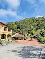 A scenic view of a picturesque estate with a large rustic house on the left, surrounded by lush greenery and trees. The bright blue sky with white clouds adds a vibrant backdrop. In the middle, there is a traditional structure with a tiled roof, and a red brick path leads through the landscaped garden area.