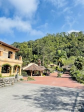 A scenic view of a picturesque estate with a large rustic house on the left, surrounded by lush greenery and trees. The bright blue sky with white clouds adds a vibrant backdrop. In the middle, there is a traditional structure with a tiled roof, and a red brick path leads through the landscaped garden area.