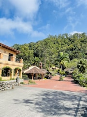 A scenic view of a picturesque estate with a large rustic house on the left, surrounded by lush greenery and trees. The bright blue sky with white clouds adds a vibrant backdrop. In the middle, there is a traditional structure with a tiled roof, and a red brick path leads through the landscaped garden area.
