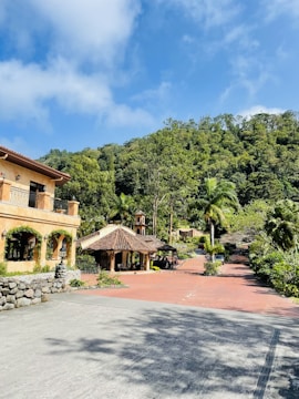 A scenic view of a picturesque estate with a large rustic house on the left, surrounded by lush greenery and trees. The bright blue sky with white clouds adds a vibrant backdrop. In the middle, there is a traditional structure with a tiled roof, and a red brick path leads through the landscaped garden area.