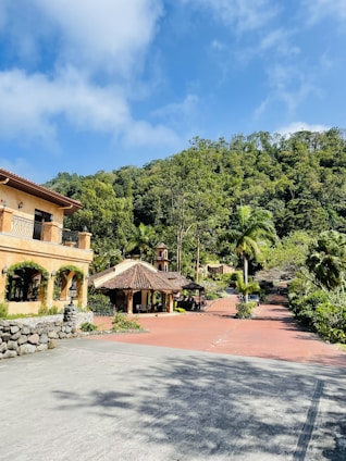 A scenic view of a picturesque estate with a large rustic house on the left, surrounded by lush greenery and trees. The bright blue sky with white clouds adds a vibrant backdrop. In the middle, there is a traditional structure with a tiled roof, and a red brick path leads through the landscaped garden area.