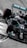 A sleek, black Formula 1 race car is taking a corner on a racetrack. The car features prominent branding and sponsorship logos, including a prominent turquoise Petronas logo. The high-speed motion is suggested by the blurred background and the angle at which the car is positioned.