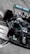 A sleek, black Formula 1 race car is taking a corner on a racetrack. The car features prominent branding and sponsorship logos, including a prominent turquoise Petronas logo. The high-speed motion is suggested by the blurred background and the angle at which the car is positioned.