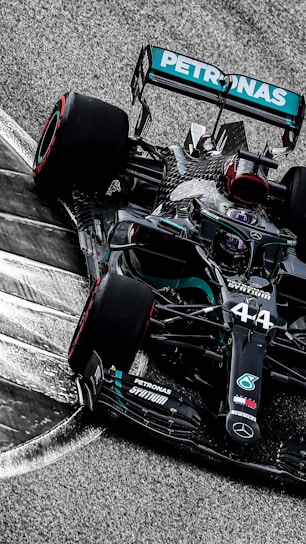 A sleek, black Formula 1 race car is taking a corner on a racetrack. The car features prominent branding and sponsorship logos, including a prominent turquoise Petronas logo. The high-speed motion is suggested by the blurred background and the angle at which the car is positioned.
