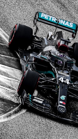 A sleek, black Formula 1 race car is taking a corner on a racetrack. The car features prominent branding and sponsorship logos, including a prominent turquoise Petronas logo. The high-speed motion is suggested by the blurred background and the angle at which the car is positioned.