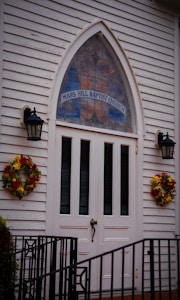 A white wooden church entrance featuring double doors with vertical panels, flanked by two black lanterns. Above the doors is a tall, arched stained glass window with 'Mars Hill Baptist Church' written on it. Two wreaths made of autumnal flowers decorate either side of the entrance. A black metal railing is visible at the bottom of the image.