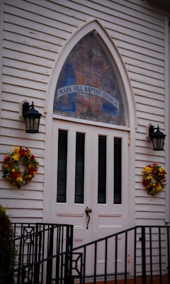 A white wooden church entrance featuring double doors with vertical panels, flanked by two black lanterns. Above the doors is a tall, arched stained glass window with 'Mars Hill Baptist Church' written on it. Two wreaths made of autumnal flowers decorate either side of the entrance. A black metal railing is visible at the bottom of the image.