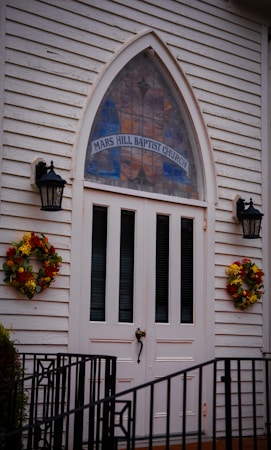 A white wooden church entrance featuring double doors with vertical panels, flanked by two black lanterns. Above the doors is a tall, arched stained glass window with 'Mars Hill Baptist Church' written on it. Two wreaths made of autumnal flowers decorate either side of the entrance. A black metal railing is visible at the bottom of the image.