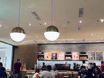 A coffee shop interior with several people seated and standing. There is a counter with baristas preparing drinks, and overhead menu boards displaying various beverage options. The lighting is warm, with large, round ceiling lights illuminating the space. The seating area has a modern and cozy design.