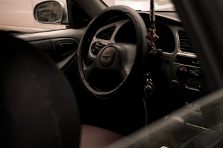 A car interior focused on the steering wheel, with a keychain hanging from it. The dashboard and control panel are visible, revealing a retro style with manual controls. The side mirror and part of the driver’s seat are also in view.