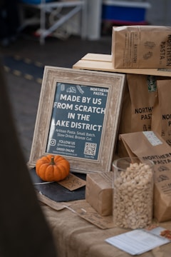 A rustic display promoting artisan pasta from the Lake District, featuring a wooden-framed sign with detailed product information. A small pumpkin adds a seasonal touch, accompanied by brown paper bags labeled with pasta types. A glass jar filled with pasta shells emphasizes the product theme.