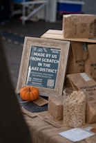 A rustic display promoting artisan pasta from the Lake District, featuring a wooden-framed sign with detailed product information. A small pumpkin adds a seasonal touch, accompanied by brown paper bags labeled with pasta types. A glass jar filled with pasta shells emphasizes the product theme.