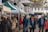 A bustling outdoor market with people walking and browsing under green and white canopy tents. Various goods such as jars and packaged items are displayed on the tables. The market is set in a quaint town environment with older-style buildings in the background.