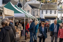 A bustling outdoor market with people walking and browsing under green and white canopy tents. Various goods such as jars and packaged items are displayed on the tables. The market is set in a quaint town environment with older-style buildings in the background.
