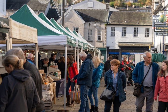 A bustling outdoor market with people walking and browsing under green and white canopy tents. Various goods such as jars and packaged items are displayed on the tables. The market is set in a quaint town environment with older-style buildings in the background.