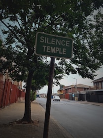 A roadside sign reading 'SILENCE TEMPLE' is prominently displayed next to a tree on a paved sidewalk. The background features a quiet street with a few parked cars and surrounding residential buildings. The scene is shaded by nearby trees, creating a tranquil atmosphere.