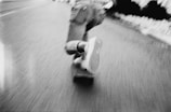 A person is riding a skateboard on a road, captured in motion. The image is in black and white, with a focus on the skateboard and shoes, creating a sense of speed and movement.