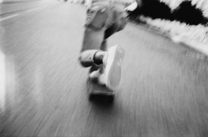 A person is riding a skateboard on a road, captured in motion. The image is in black and white, with a focus on the skateboard and shoes, creating a sense of speed and movement.