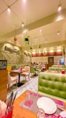 An indoor dining area with modern decor, featuring green upholstered seating booths, tables set with plates and glasses, and bright overhead lighting. Hanging plants are suspended from the ceiling, and there are geometric patterns on the walls.