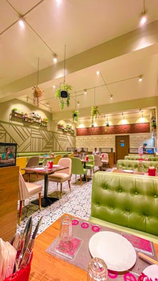 An indoor dining area with modern decor, featuring green upholstered seating booths, tables set with plates and glasses, and bright overhead lighting. Hanging plants are suspended from the ceiling, and there are geometric patterns on the walls.