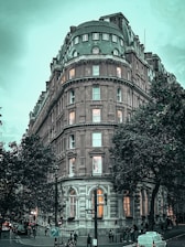 A majestic, historic hotel building with ornate architectural details, situated at a street corner. The building features a rounded facade with multiple lit windows and a grand entrance. Trees surround the area, and people are walking on the street with vehicles parked nearby.