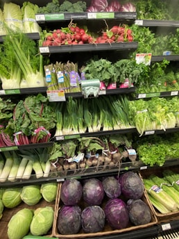 A well-stocked supermarket produce section displays a variety of fresh vegetables, including radishes, asparagus, kale, and cabbages. The produce is neatly organized on shelves, with vibrant greens and purples dominating the colors.