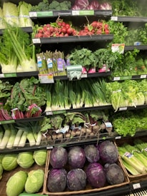 A well-stocked supermarket produce section displays a variety of fresh vegetables, including radishes, asparagus, kale, and cabbages. The produce is neatly organized on shelves, with vibrant greens and purples dominating the colors.