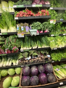 A well-stocked supermarket produce section displays a variety of fresh vegetables, including radishes, asparagus, kale, and cabbages. The produce is neatly organized on shelves, with vibrant greens and purples dominating the colors.