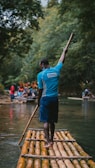 A person stands on a bamboo raft holding a long stick, surrounded by lush green trees. Other people and rafts are visible in the background, creating an adventurous and serene natural setting.