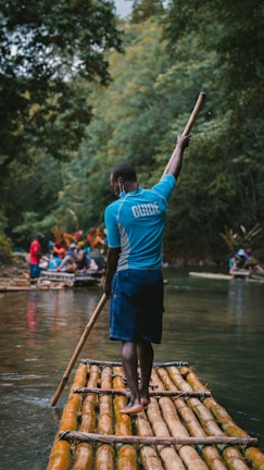 A person stands on a bamboo raft holding a long stick, surrounded by lush green trees. Other people and rafts are visible in the background, creating an adventurous and serene natural setting.