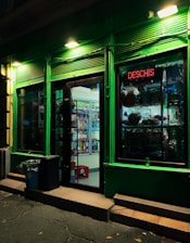 A small retail store is illuminated at night, with a glowing green exterior and a red neon sign that says 'DESCHIS'. The interior is visible through the glass door, showing shelves stocked with a variety of goods, including drinks and food items. There is a trash bin and a step leading to the entrance.
