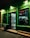 A small retail store is illuminated at night, with a glowing green exterior and a red neon sign that says 'DESCHIS'. The interior is visible through the glass door, showing shelves stocked with a variety of goods, including drinks and food items. There is a trash bin and a step leading to the entrance.