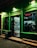 A small retail store is illuminated at night, with a glowing green exterior and a red neon sign that says 'DESCHIS'. The interior is visible through the glass door, showing shelves stocked with a variety of goods, including drinks and food items. There is a trash bin and a step leading to the entrance.