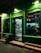 A small retail store is illuminated at night, with a glowing green exterior and a red neon sign that says 'DESCHIS'. The interior is visible through the glass door, showing shelves stocked with a variety of goods, including drinks and food items. There is a trash bin and a step leading to the entrance.
