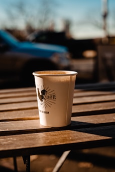 A disposable coffee cup sits on a wooden table under warm sunlight. The cup features a rooster logo and the word 'coffee' printed on it. In the background, there is a blurred view of a car and trees, suggesting an outdoor setting.