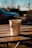 A disposable coffee cup sits on a wooden table under warm sunlight. The cup features a rooster logo and the word 'coffee' printed on it. In the background, there is a blurred view of a car and trees, suggesting an outdoor setting.