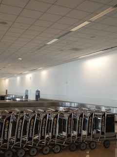 A row of luggage carts is neatly lined up next to an empty baggage claim conveyor belt in a large, spacious airport terminal. The ceiling features a grid pattern with recessed lighting, and the walls are plain and light-colored.
