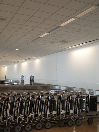A row of luggage carts is neatly lined up next to an empty baggage claim conveyor belt in a large, spacious airport terminal. The ceiling features a grid pattern with recessed lighting, and the walls are plain and light-colored.