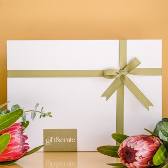 A white gift box tied with an olive green ribbon is centrally displayed against a muted mustard background. The box is accompanied by two pink protea flowers and green foliage at the bottom corners. A small card with the words 'gift&crate' is placed below the box.