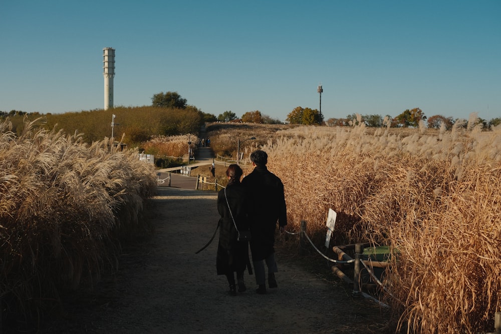 a man and woman walking down a dirt road with tall grass on either side of them