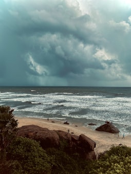 A beach scene features overcast skies with dramatic, swirling clouds suggesting an impending storm. The ocean has moderate waves crashing onto a sandy beach. Several people are scattered along the shoreline and atop rocks, with lush green vegetation in the foreground.