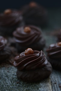 Dark chocolate desserts are arranged a textured wooden surface. The confectionery features swirled chocolate frosting with a small brown topping on each piece. The atmosphere is dimly lit, emphasizing the rich texture and color of the chocolate.