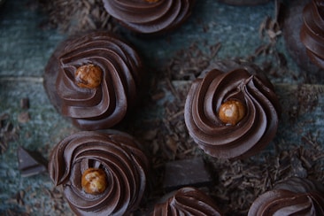 Rich chocolate cupcakes with swirled frosting topped with hazelnuts are scattered on a rustic wooden surface surrounded by chocolate shavings.