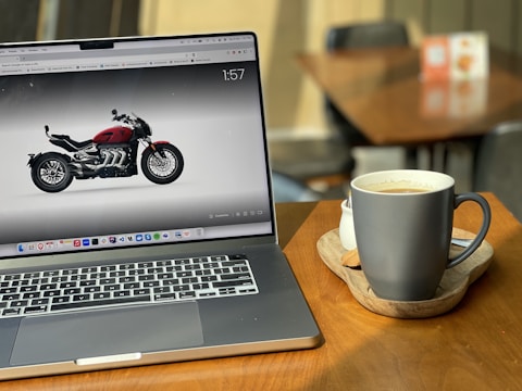 A laptop displaying an image of a motorcycle is situated on a wooden table. Beside the laptop is a cup of coffee on a wooden tray. The scene takes place in a cozy setting with a blurred background, which includes chairs and a table.