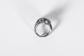 A detailed and intricate silver ring with a textured design, resembling an ornate piece possibly inspired by nature or historical motifs. It casts a soft shadow on a plain white background, highlighting its craftsmanship.