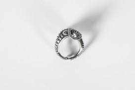 A detailed and intricate silver ring with a textured design, resembling an ornate piece possibly inspired by nature or historical motifs. It casts a soft shadow on a plain white background, highlighting its craftsmanship.