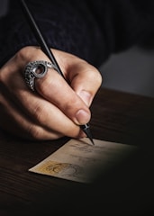A close-up of a hand holding a black pen, writing on a vintage postcard. The hand wears a distinctive ring shaped like an octopus tentacle, adding an artistic touch. The postcard features elegant cursive handwriting and an old stamp with a circular postmark in the corner. The background is a dark wooden surface, enhancing the focus on the hand and postcard.